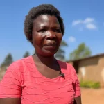 Dorothy Masasa(in a pink blouse )standing on a compound with a house in the background.