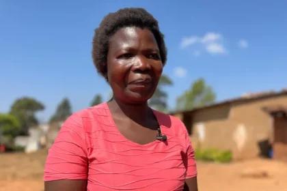 Dorothy Masasa(in a pink blouse )standing on a compound with a house in the background.