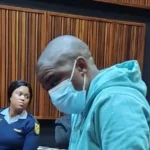 Nkosinathi Phakathi in a courtroom wearing a nose mask surrounded by police officers.