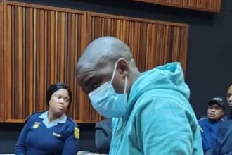 Nkosinathi Phakathi in a courtroom wearing a nose mask surrounded by police officers.