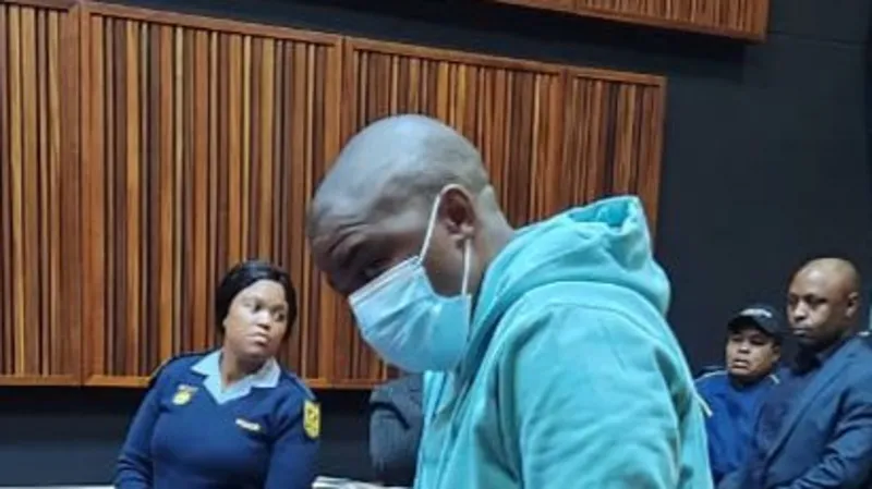 Nkosinathi Phakathi in a courtroom wearing a nose mask surrounded by police officers.