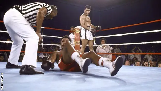 Muhammad Ali watches on as George Foreman struggles to get up after being knocked out.